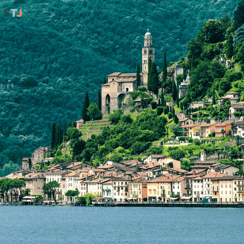 Landscape of the canton of Ticino, in Italian-speaking Switzerland, where a lakeside village and a hillside church reflect the blend of Alpine identity and Mediterranean aesthetics.