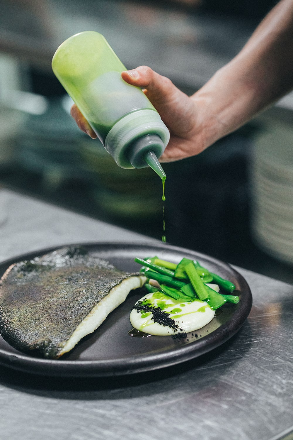 Mano sirviendo una salsa verde sobre un plato de pescado en una cocina profesional.