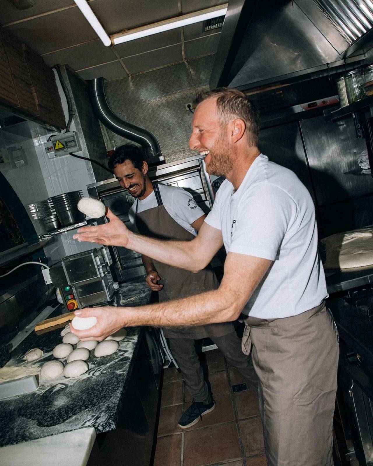 Dos cocineros trabajando masa en una cocina profesional, fotografiados con flash en un momento de movimiento y complicidad.