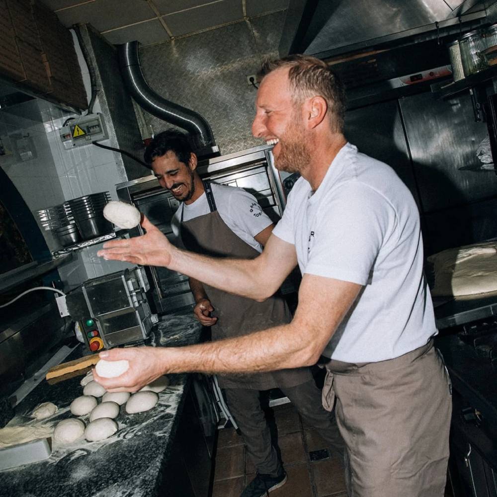 Fotografía gastronómica de cocina profesional con flash, harina en el aire y cocineros trabajando la masa.