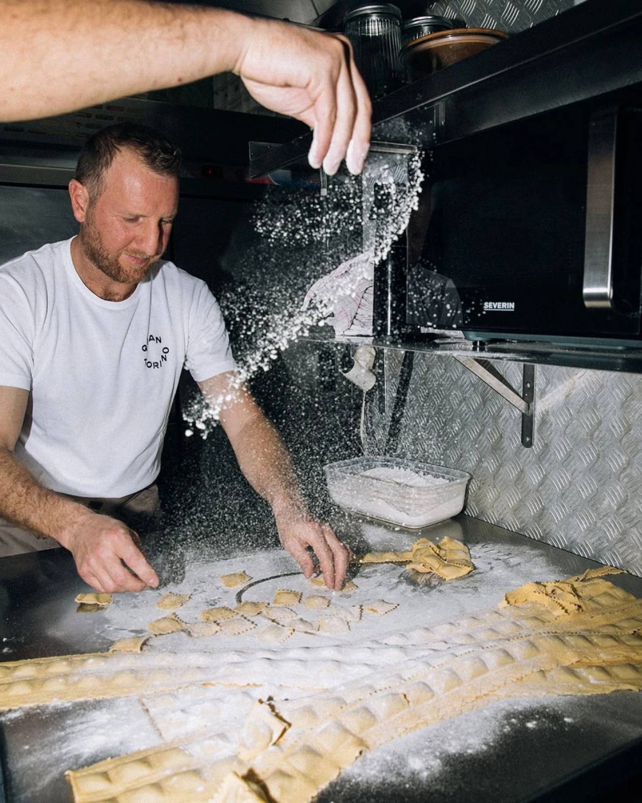 Cocinero preparando pasta fresca en una cocina profesional con harina en el aire, fotografiado con flash.