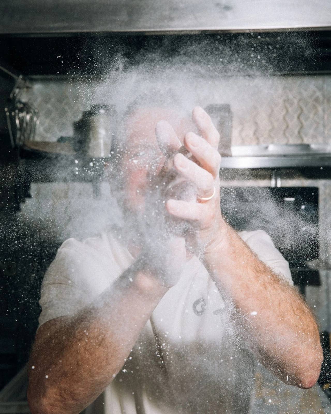 Cocinero en una cocina profesional rodeado de harina en suspensión, fotografiado con flash durante la preparación de pasta.
